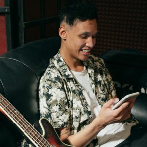 Smiling man with electric guitar checking his smartphone while sitting comfortably on a sofa.