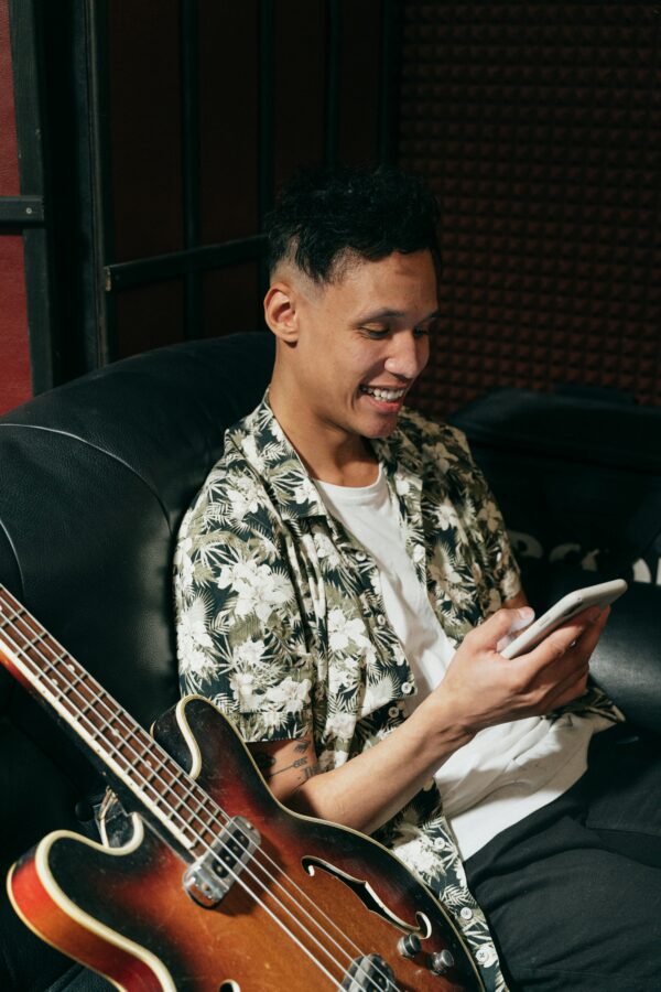 Smiling man with electric guitar checking his smartphone while sitting comfortably on a sofa.
